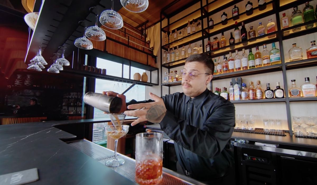 A bartender prepares a drink at a bar, with bottles and cocktail shakers around him.
