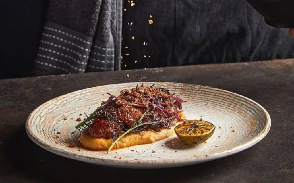 Un plato con un plato de carne mientras una persona le añade sal, destacando técnicas exclusivas de autor en La Makha