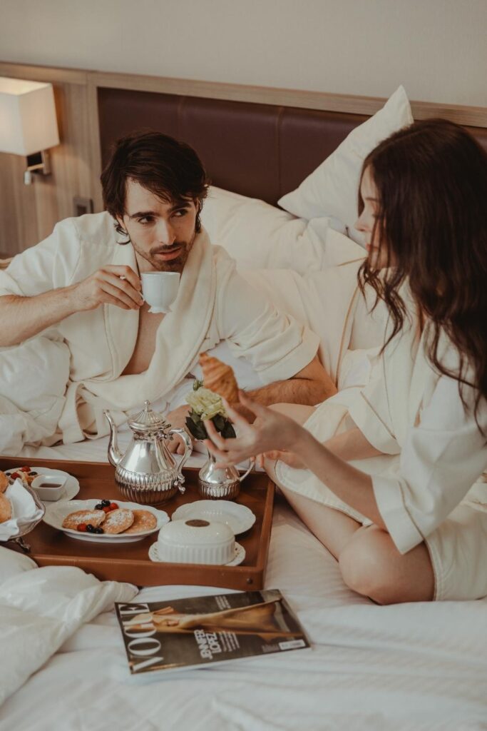 Man and woman in bathrobes, sitting on bed, enjoying a meal together in a romantic setting