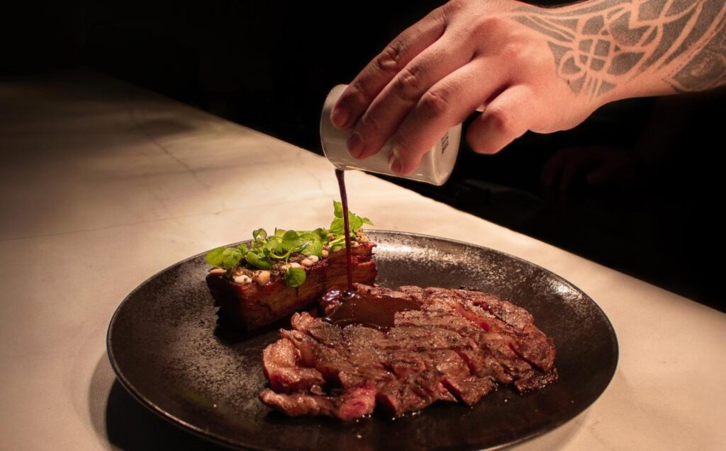 una persona vierte salsa sobre un jugoso filete en un plato, resaltando su presentación deliciosa.