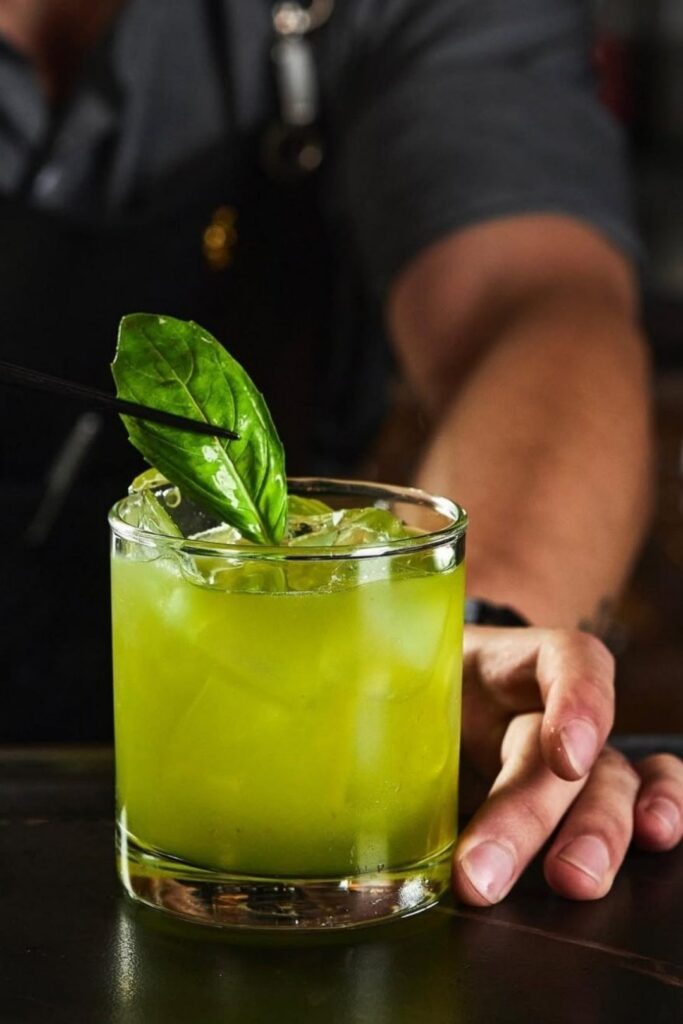 Bartender sosteniendo un trago verde decorado, invitando a disfrutar de La Makha