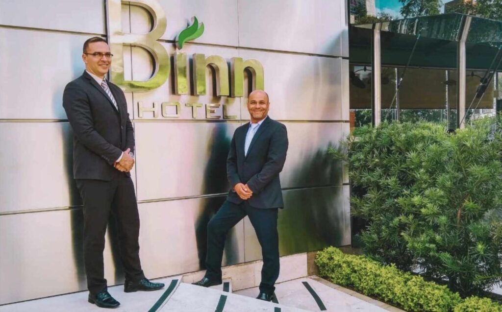 Dos hombres en trajes de pie frente al hotel Binn, sonriendo y conversando.