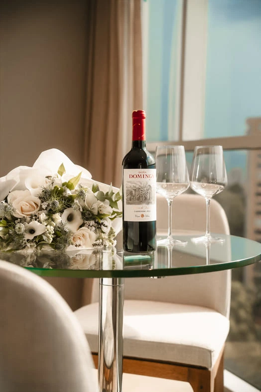 Wine bottle, two glasses and flower bouquet on glass table with balcony view.