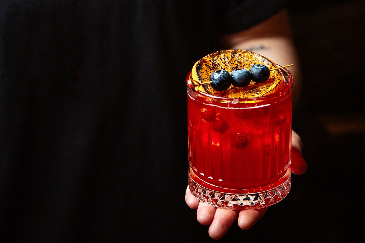 Experiencia de coctelería artesanal en La Makha Medellín: Bartender sosteniendo un cóctel vibrante de frutos rojos con guarnición de naranja quemada y arándanos frescos.