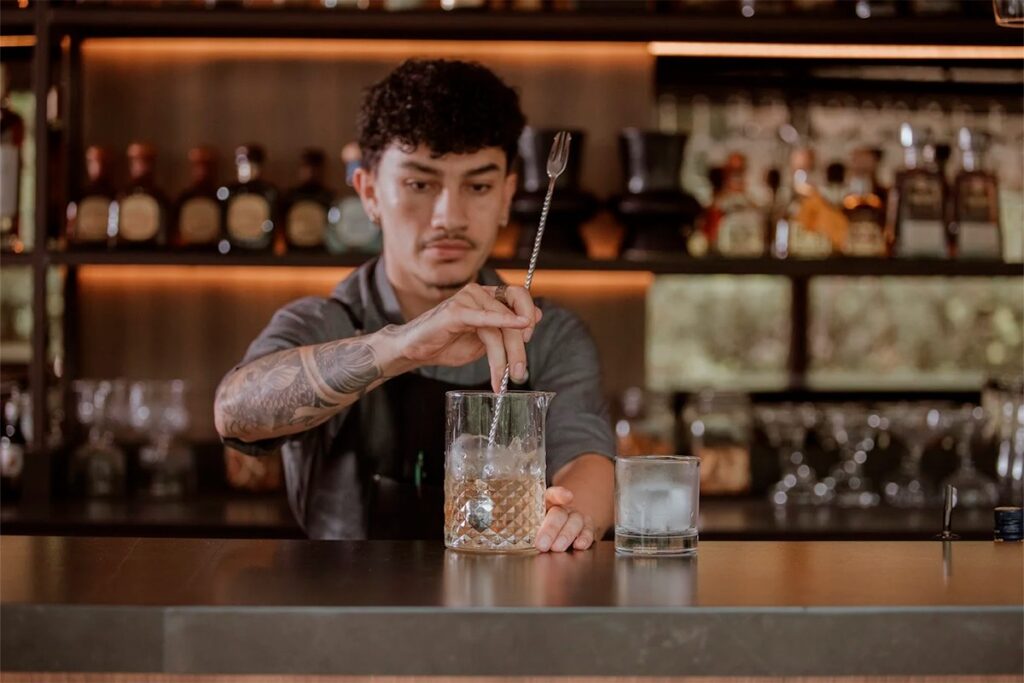 Experiencia de coctelería artesanal en La Makha Medellín: Bartender con tatuajes removiendo con elegancia un cóctel premium en un vaso de mezcla cristalino.