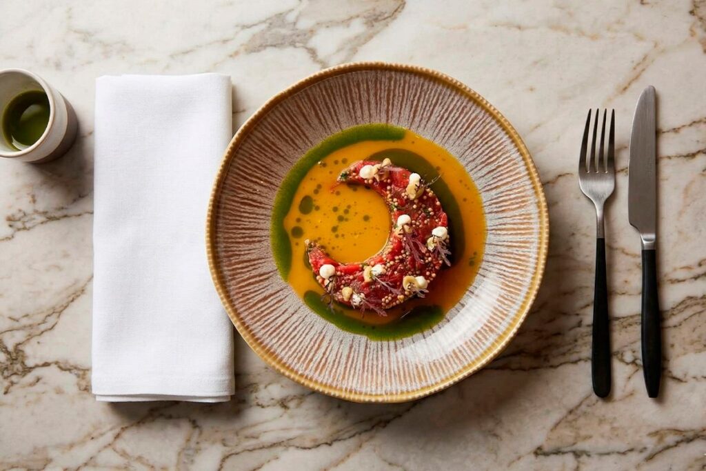 Plato insignia de cocina fusión en La Makha Medellín: Carpaccio de res en forma de media luna con emulsión cítrica y aceites herbales sobre mesa de mármol.