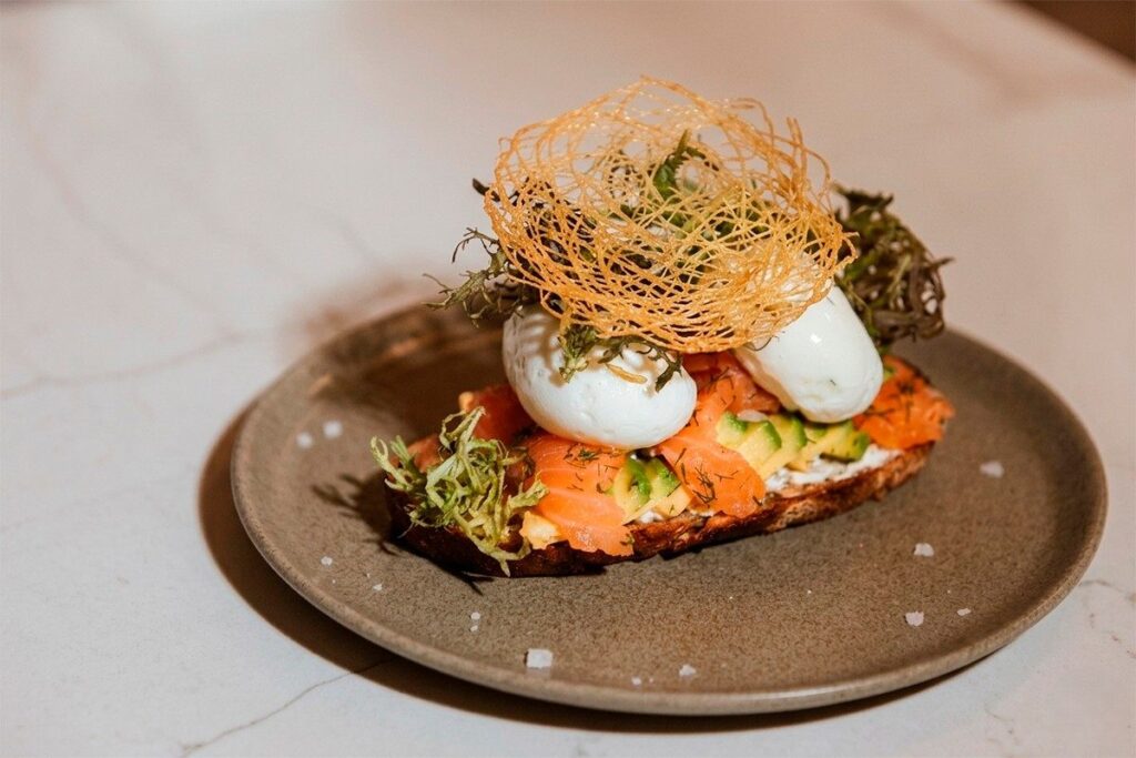 Tostada gourmet con salmón, aguacate y huevos pochados, decorada con crujiente artesanal en un plato de alta cocina