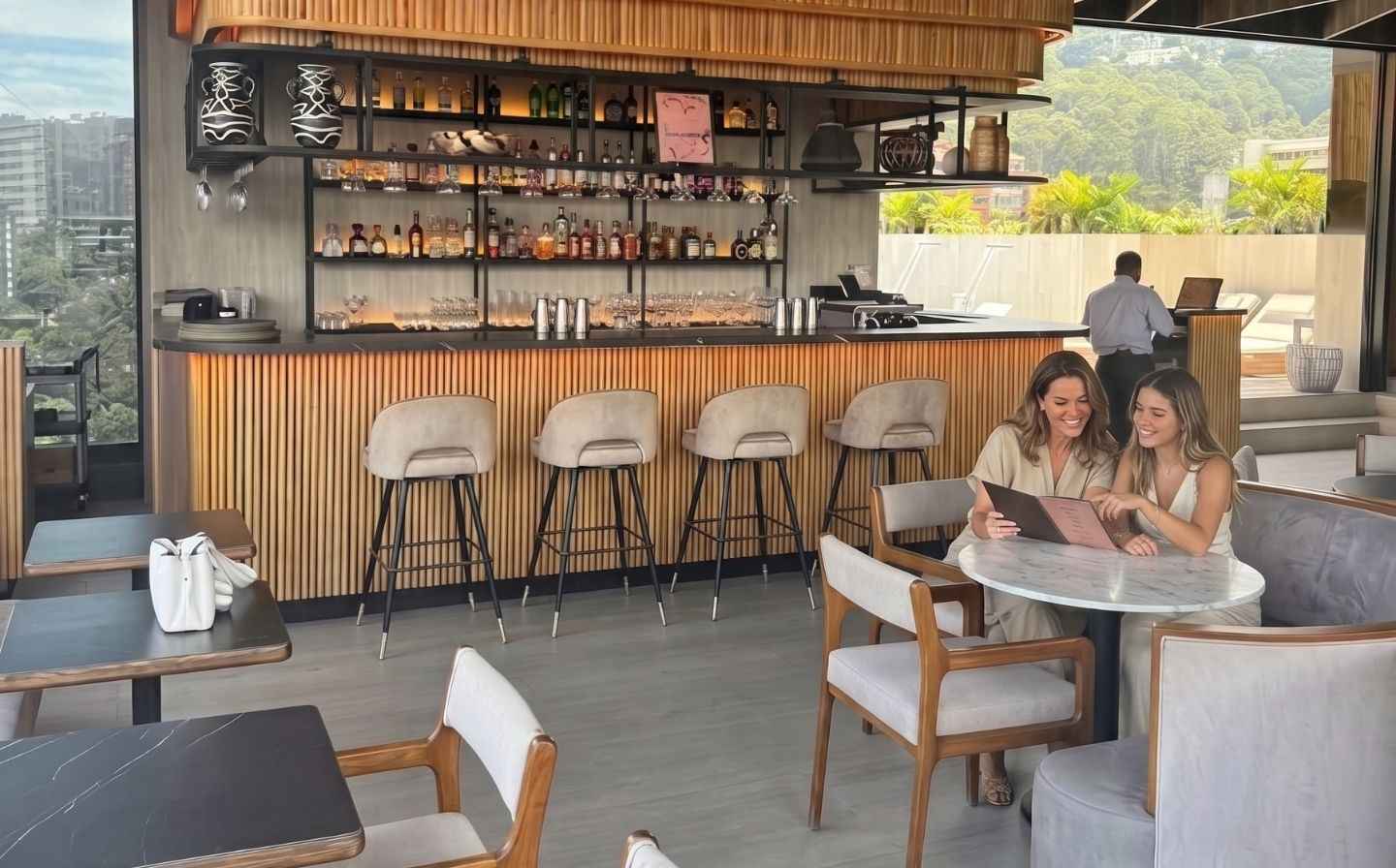 Madre e hija sonriendo mientras ven el menú en Etro Rooftop Medellín, con barra de madera moderna y vista a la ciudad.