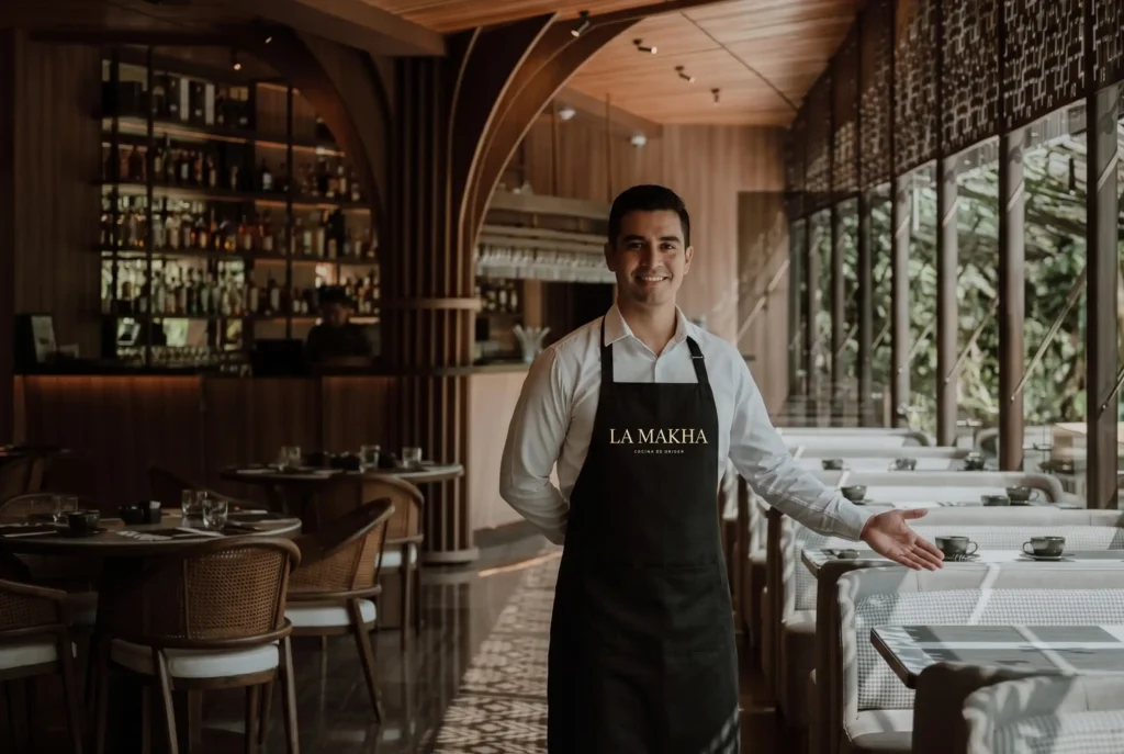 Mesero sonriente en el restaurante La Makha, referente de fine dining Medellín, con diseño interior en madera y bar al fondo.