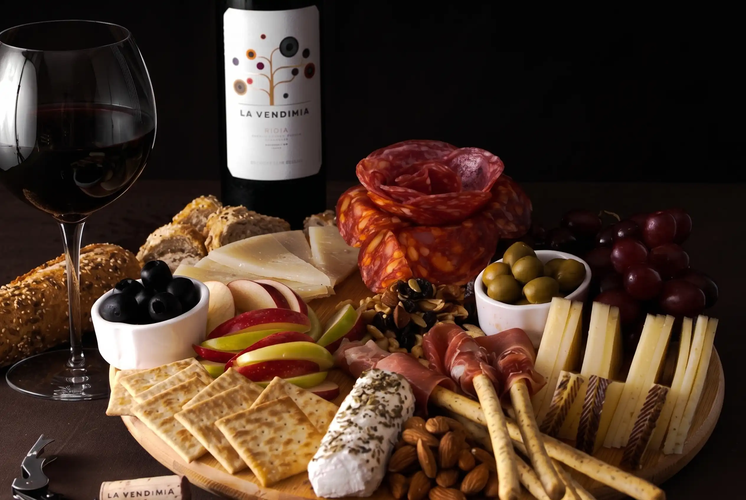 Tabla de quesos maduros, carnes frías y frutos secos junto a una copa y botella de vino tinto para maridaje.