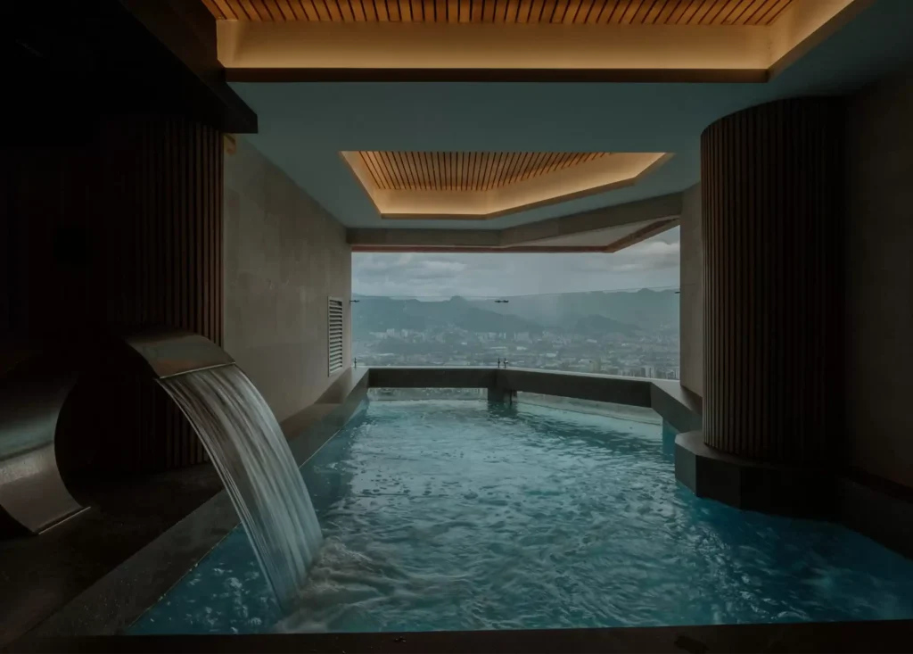 Piscina interior techada con una cascada de metal, iluminación cálida en el techo y un gran ventanal con vista a la ciudad.