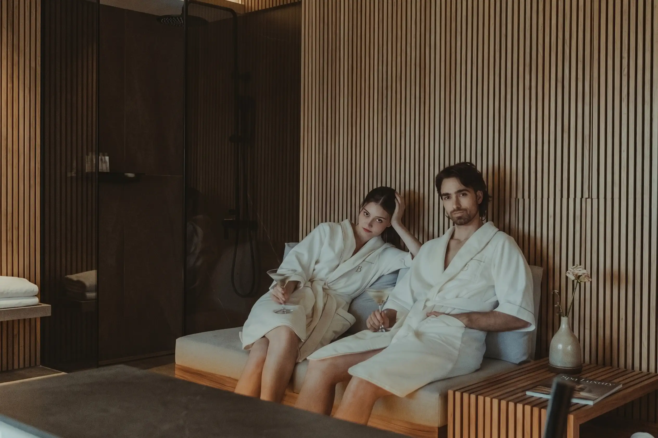 Pareja en batas de spa de BINN Hotel descansando con copas en sala de bienestar en madera