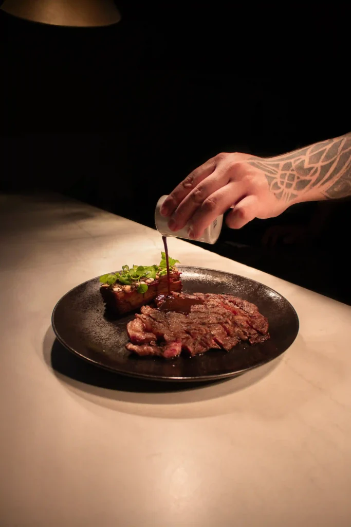 Chef sirviendo salsa en corte de carne laminado con costilla y microgreens en La Makha Medellín
