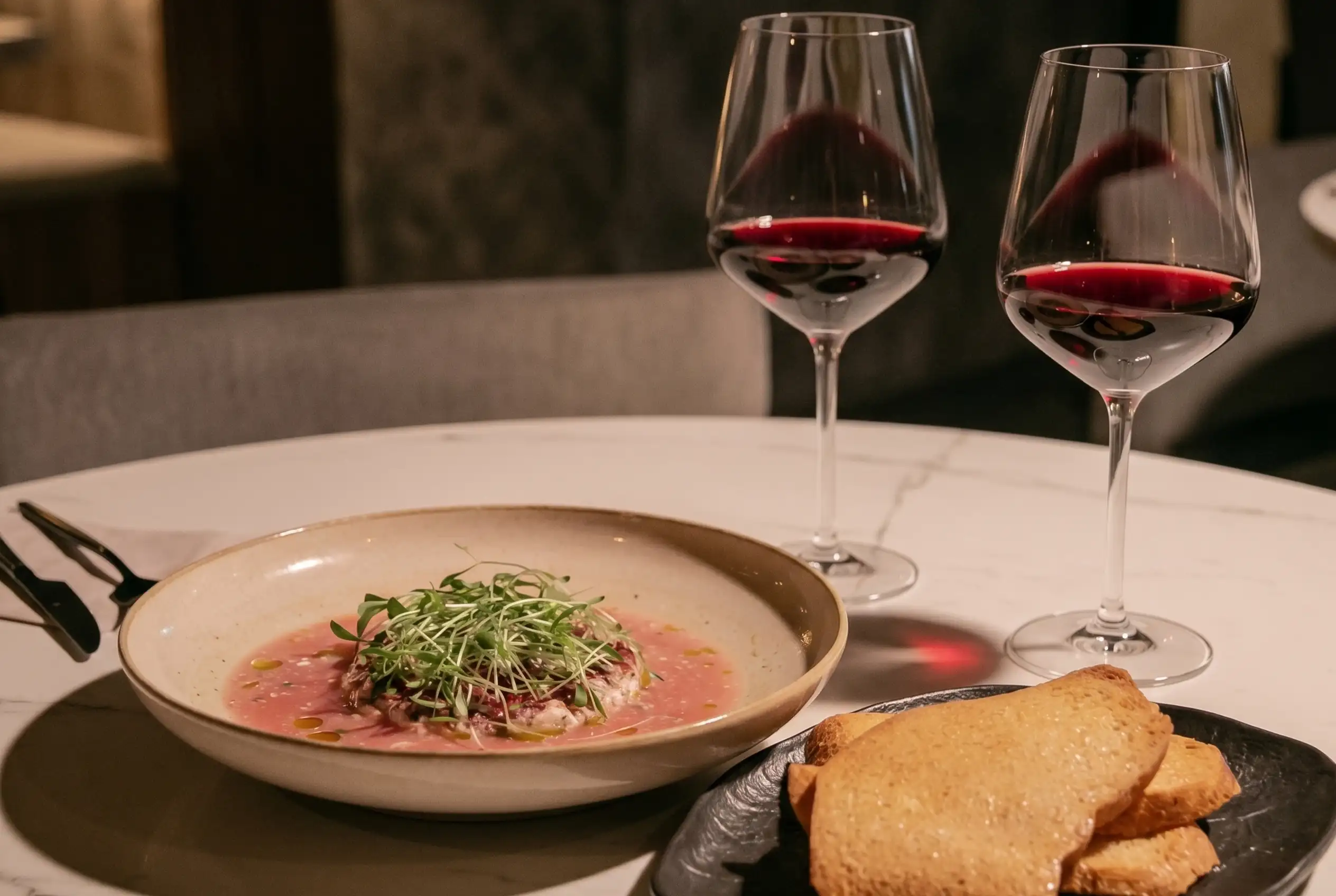 Carpaccio con microgreens y dos copas de vino tinto sobre mesa de mármol en La Makha Medellín