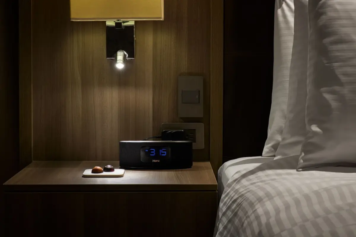 Mesa de noche de madera en Binn Hotel Medellín con reloj despertador digital, lámpara de lectura y chocolates de bienvenida junto a la cama