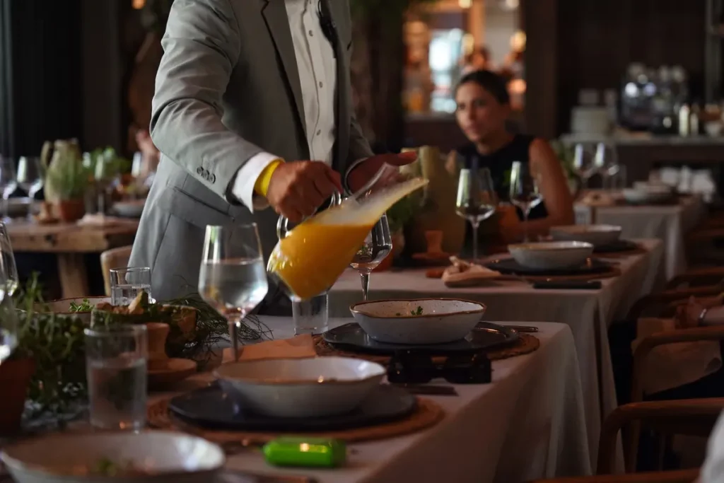 Mesero con traje gris sirviendo una bebida en jarra sobre una mesa elegante en el restaurante La Makha de Medellín.