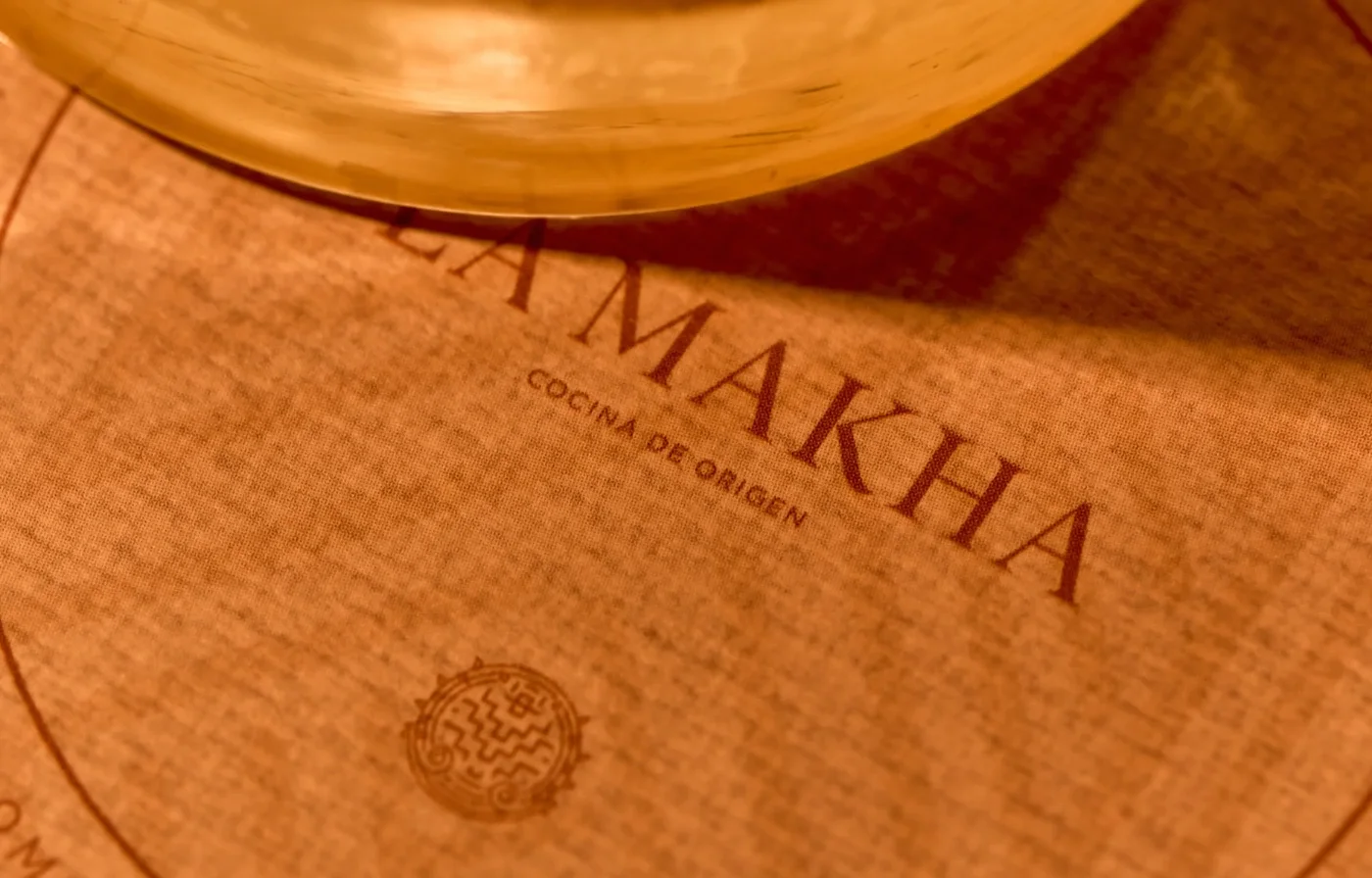 Detalle del logo y menú de cocina de origen para el desayuno en La Makha Medellín sobre un mantel individual con iluminación cálida.