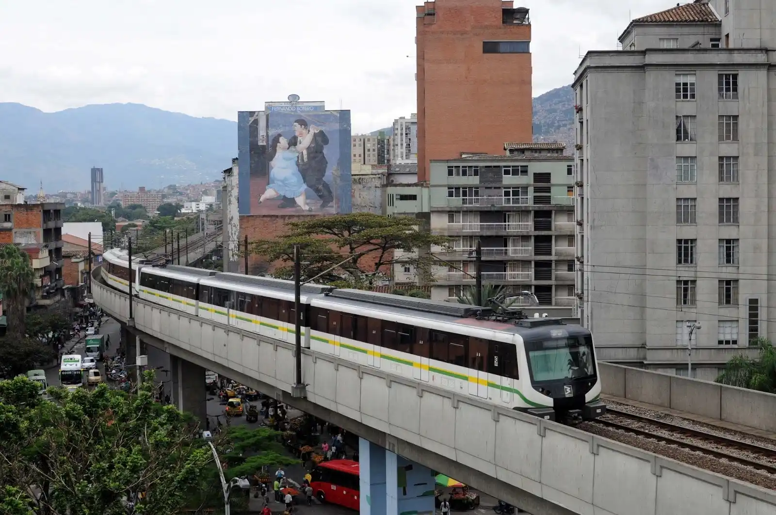 Tren del Metro de Medellín transitando en viaducto elevado frente a edificios del centro y un mural de Fernando Botero al fondo.