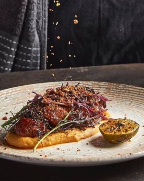 Un plato con un plato de carne mientras una persona le añade sal, destacando técnicas exclusivas de autor en La Makha