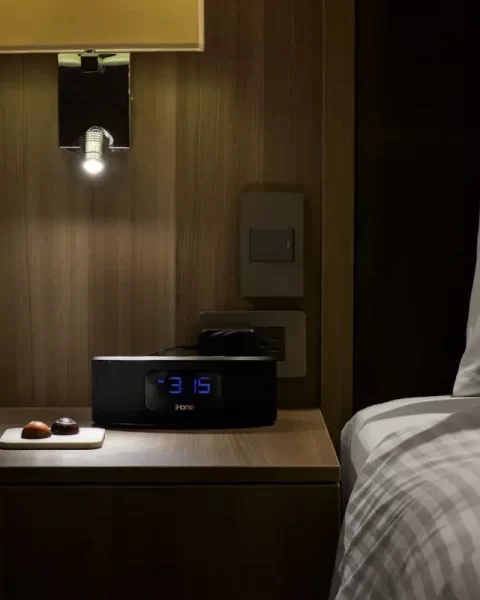 Mesa de noche de madera en Binn Hotel Medellín con reloj despertador digital, lámpara de lectura y chocolates de bienvenida junto a la cama