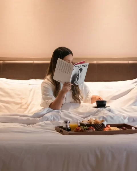 Persona leyendo en cama de BINN Hotel con bandeja de desayuno y ropa de cama blanca.
