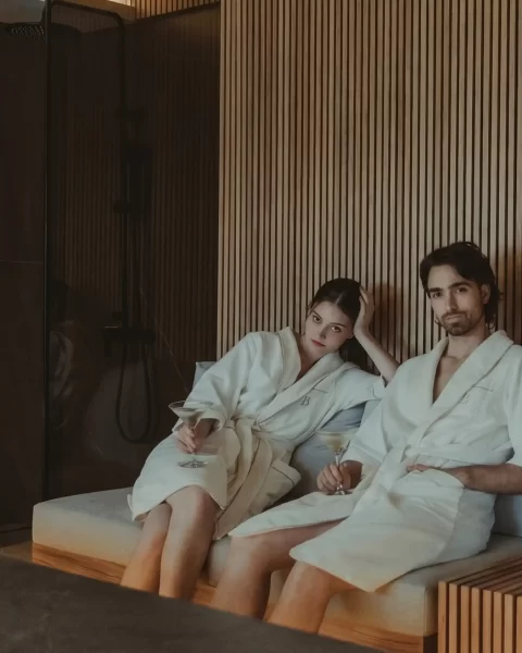 Pareja en batas de spa de BINN Hotel descansando con copas en sala de bienestar en madera