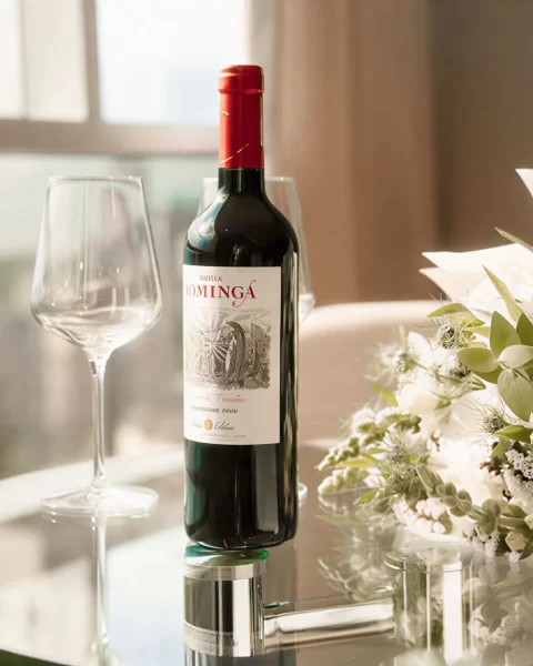 Botella de vino, copa y ramo de flores sobre mesa de vidrio en habitación con vista.