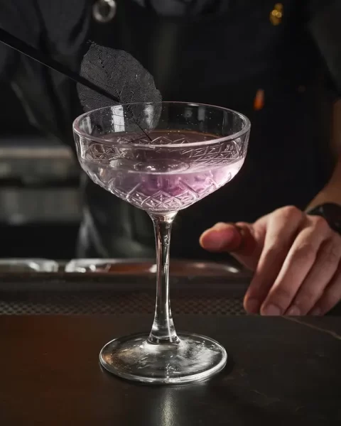 Bartender decorando un cóctel artesanal de color violeta con una hoja seca en la barra de La Makha en Medellín.