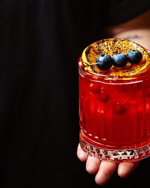 Experiencia de coctelería artesanal en La Makha Medellín: Bartender sosteniendo un cóctel vibrante de frutos rojos con guarnición de naranja quemada y arándanos frescos.