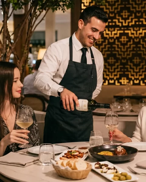 Mesero sirviendo vino blanco a dos mujeres en un restaurante de fine dining Medellín con ambiente elegante y moderno.