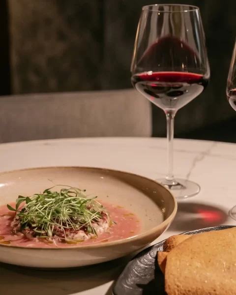 Carpaccio con microgreens y dos copas de vino tinto sobre mesa de mármol en La Makha Medellín