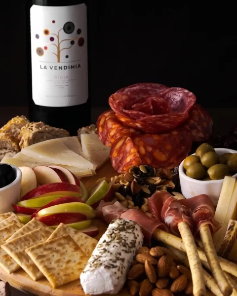 Tabla de quesos maduros, carnes frías y frutos secos junto a una copa y botella de vino tinto para maridaje.