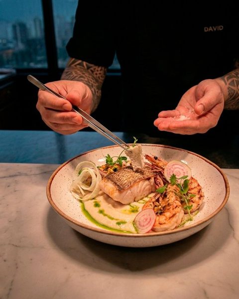 Chef en La Makha Medellín finalizando un plato del menú de temporada de mayo con pinzas de precisión y una vista panorámica de la ciudad al fondo.