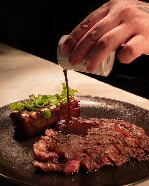 una persona vierte salsa sobre un jugoso filete en un plato, resaltando su presentación deliciosa.